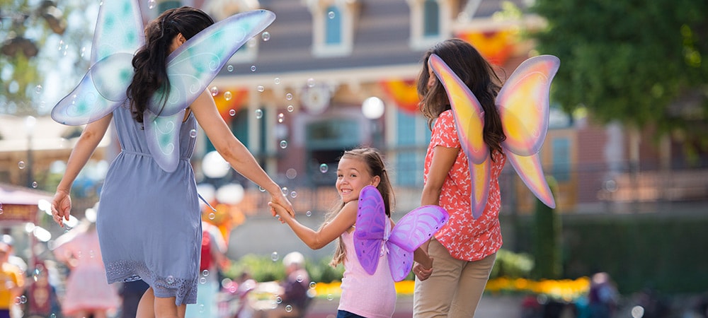 Una mamá, su hija y una amiguita caminan juntas por el parque Disneyland con sus alas de hada.