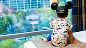 A young boy wearing a Mickey Mouse ear hat sits on a bed inside his hotel room while glancing out the window and pointing at the Disneyland Hotel pool area A young boy wearing a Mickey Mouse ear hat sits on a bed inside his hotel room while glancing out the window and pointing at the Disneyland Hotel pool area