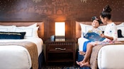 Two children read a book while sitting side by side on a bed inside of their hotel room