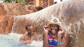 A man and a woman in a hat, laugh while under a waterfall in a pool at Aulani Resort A man and a woman in a hat, laugh while under a waterfall in a pool at Aulani Resort