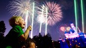 A excited young boy sits on his dad's shoulders and points to the exploding Disneyland Park fireworks