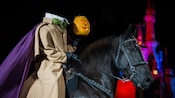 A headless horseman rides a horse and holds a candlelit jack-o’-lantern near Cinderella Castle
