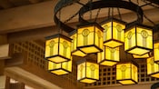 A chandelier suspended from a wood bean ceiling consists of several identical frosted glass boxes designed in a geometric pattern that hang by chains from concentric iron hoops