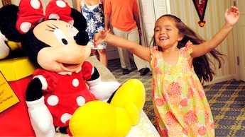 A small girl in a hotel guest room with her parents is overjoyed to find a large Minnie Mouse plush on the bed