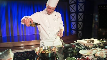 A chef in a demonstration kitchen pours oil into a sautÃ© pan containing scallops and vegetables