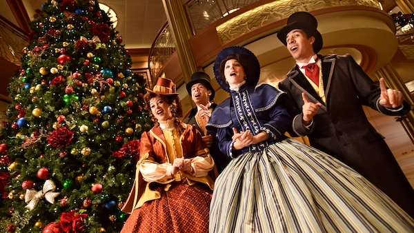 4 carolers dressed in 19th century attire singing in front of a Christmas tree