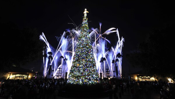 A tree decorated with Christmas lights and topped by an angel near a crowd of people, trees and fireworks