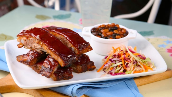 A plate of Spice-rubbed pork spare ribs with baked beans and cole slaw sits on an outdoor café table at the River Belle Restaurant