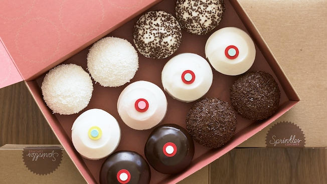 One dozen cupcakes with a variety of decorations and toppings in an open cardboard box