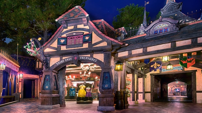 A shop with a half-timber style facade, a sign that reads 'Fairy Tale Treasure' and merchandise that includes ornate princess dresses for children, next to a tunnel with a sign that reads 'To Frontierland'