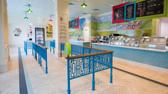 An ice cream parlor with an intricately patterned iron and wood railing to control customer flow in the waiting area, a freezer and display cases to one side, 2 long ice cream serving counters with tile and glass fronts and signage featuring their menu hanging from the ceiling