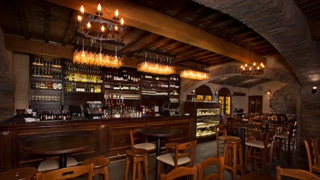 A dark-wood and brick wine cellar with high tables, stools, and a long bar with a wall of wine bottles