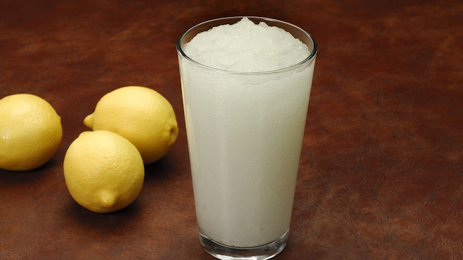 A glass of frozen lemonade sits on a table next to 3 lemons