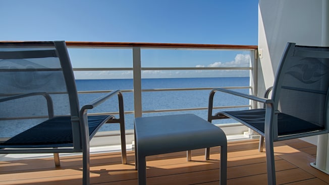 Two deck chairs flank a low table in front of a railing with a partially obstructed ocean view