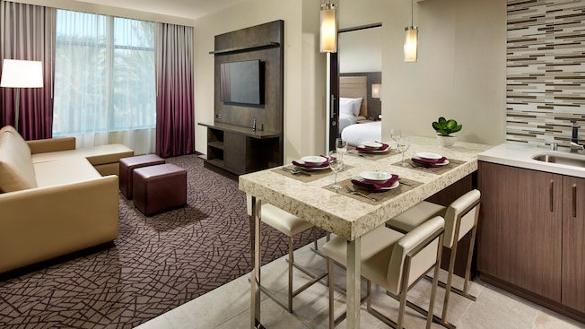 otels near Disneyland for large families: Living room has dining table and 4 chairs, sink, sofa with lamp, upholstered ottomans and entertainment center, and beyond, the bedroom