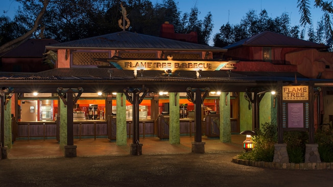 The Flame Tree Barbecue housed in a traditional African style building with an outdoor service area, consisting of a patio sheltered by a corrugated tin roof, a sign with the restaurant name on top of the roof and a menu board located to the right side as you approach