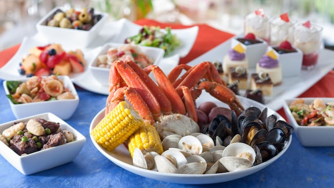 Plate of crab legs, clams, muscles, corn on the cob and potatoes, next to other plates of food