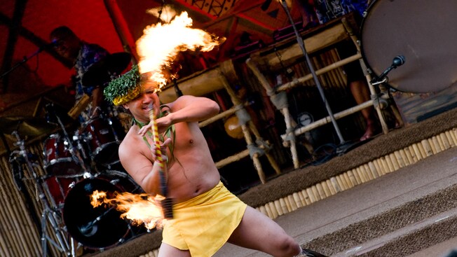 A shirtless Cast Member in a Polynesian outfit dancing and twirling a flaming baton