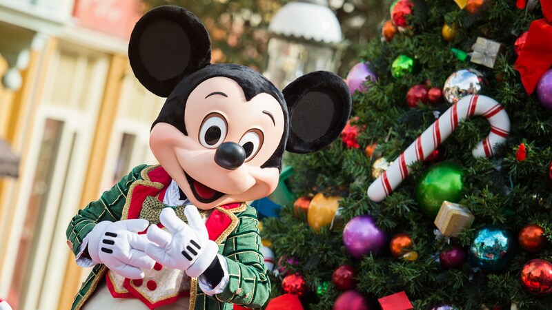 Mickey Mouse dressed in festive holiday clothing while standing next to a decorated Christmas tree