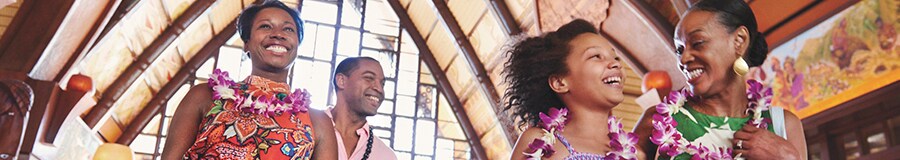 A multi-generational family smile and laugh as they walk through the main lobby at Aulani Resort wearing flower and kukui nut leis