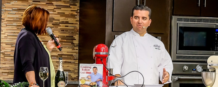 A woman with a microphone speaking to Chef Buddy Valastro in a demonstration kitchen containing a mixing machine and one of the chefâs cookbooks